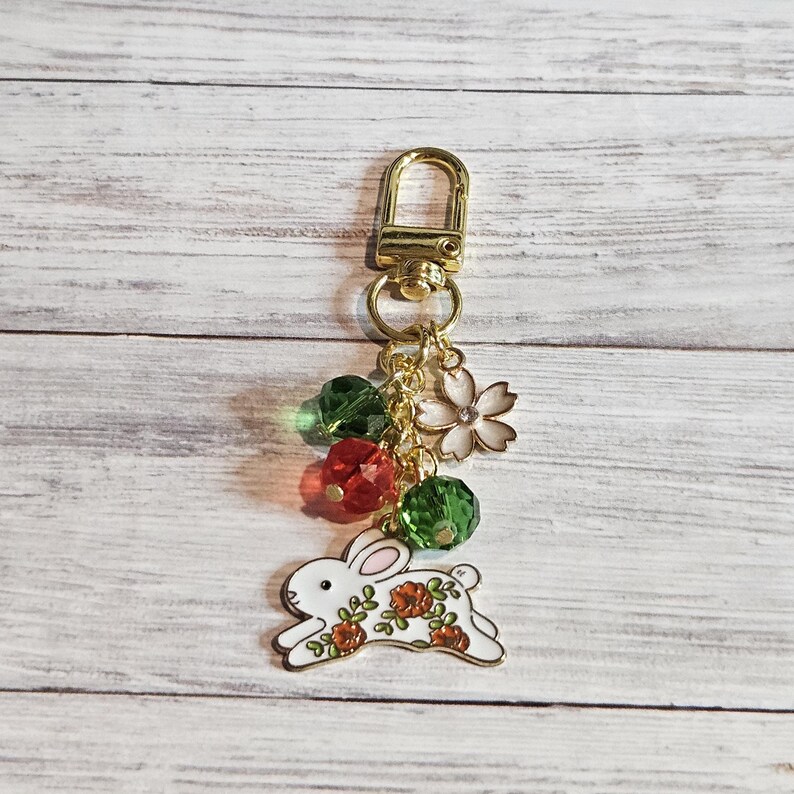 May include: A gold-colored keychain with a white rabbit charm decorated with red and orange flowers. The keychain also features green and red crystal beads, a white flower charm, and a gold clasp.