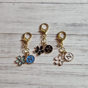 May include: Three gold-coloured keychains with charms. Each charm features a flower and a circular pendant with the letter "N". The flowers are in blue, black, and white. The pendants are in blue, black, and white.