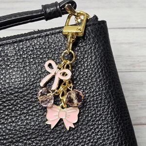 May include: A gold-toned charm with pink bow and crystal accents, attached to a black leather bag. The charm features a small bow and two crystal beads, adding a touch of elegance to the accessory.