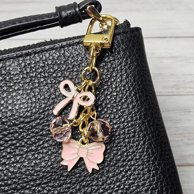 May include: A gold-toned charm with pink bow and crystal accents, attached to a black leather bag. The charm features a small bow and two crystal beads, adding a touch of elegance to the accessory.