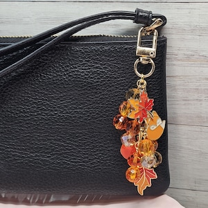 May include: A black textured wristlet bag featuring a gold-toned metal lobster clasp keychain. The keychain is adorned with a cluster of orange and yellow faceted crystal beads, a small orange fox charm, and two maple leaf charms in red and orange.