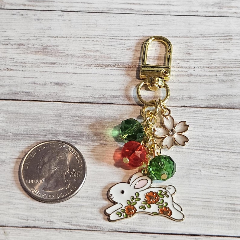 May include: A gold-colored keychain with a white rabbit charm decorated with orange flowers. The keychain also features green and red faceted beads, a white flower charm, and a gold clasp. A quarter is next to the keychain.