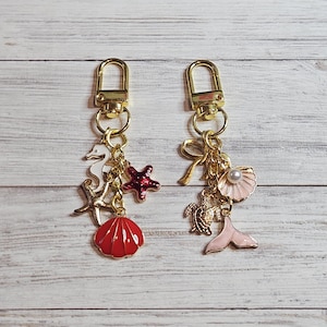 May include: Two gold-toned keychains with sea-themed charms. One keychain features a seahorse, starfish, and a red seashell. The other has a bow, a pearl, a seashell, a turtle, and a pink mermaid tail. The charms are in various colors.