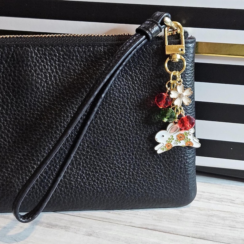 May include: Black leather wristlet with a gold-tone zipper and wrist strap. A decorative charm hangs from the zipper, featuring a white bunny with orange floral accents, red and green beads, and a white flower.