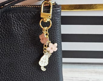 May include: A gold-colored keychain with a black leather wallet. The keychain features a cat charm with pink flower accents. The cat charm is white with gray details. The flowers are a light pink color.