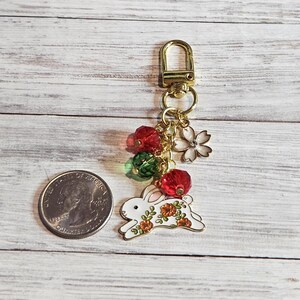 May include: A gold-toned keychain with a white bunny charm decorated with orange flowers. The charm is accented with red and green beads, a white flower, and a gold clasp. The keychain is shown next to a U.S. quarter.