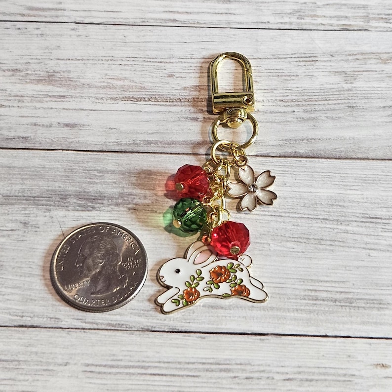 May include: A gold-toned keychain with a white bunny charm decorated with orange flowers. The charm is accented with red and green beads, a white flower, and a gold clasp. The keychain is shown next to a U.S. quarter.