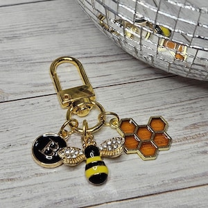 May include: Gold-toned keychain with bee and honeycomb charms. The bee charm is black and yellow with rhinestone wings. A black circular charm with the letter "B" is also included. The honeycomb charm is orange and gold.