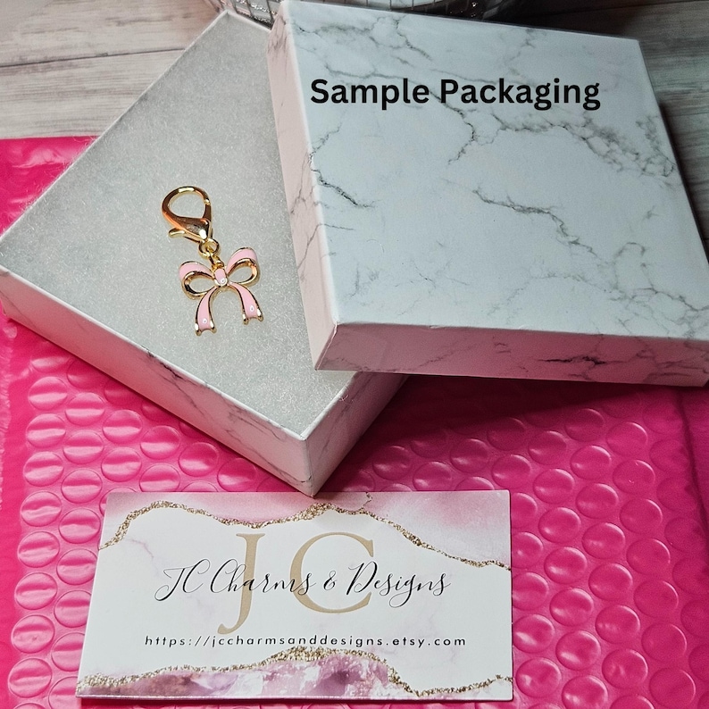 May include: A gold-colored charm with a pink bow design, presented in a marble-patterned box. The box is open, revealing the charm. A business card with the text "JC Charms & Designs" is also visible.