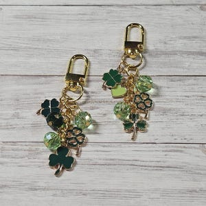 May include: Two gold-toned metal keychains featuring dangling charms of green enamel shamrocks and four-leaf clovers, interspersed with faceted light green and dark green glass beads, all attached to a gold lobster claw clasp.