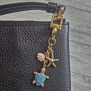 May include: A gold-colored charm with a blue and gold turtle, a white seashell, and a gold starfish hanging from a gold clasp. The charm is attached to a black leather purse.