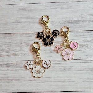 May include: Three gold-coloured keychains with lobster claw clasps. Each keychain features a flower charm and a circular charm with a letter. One keychain has black flowers and a black letter Z. Another has pink flowers and a pink letter J. The last has white flowers and a white letter E.