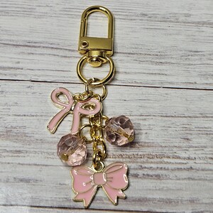 May include: A gold-colored keychain with a lobster clasp, featuring pink enamel bows and faceted pink beads. The keychain is suspended from a gold chain, creating a charming accessory.
