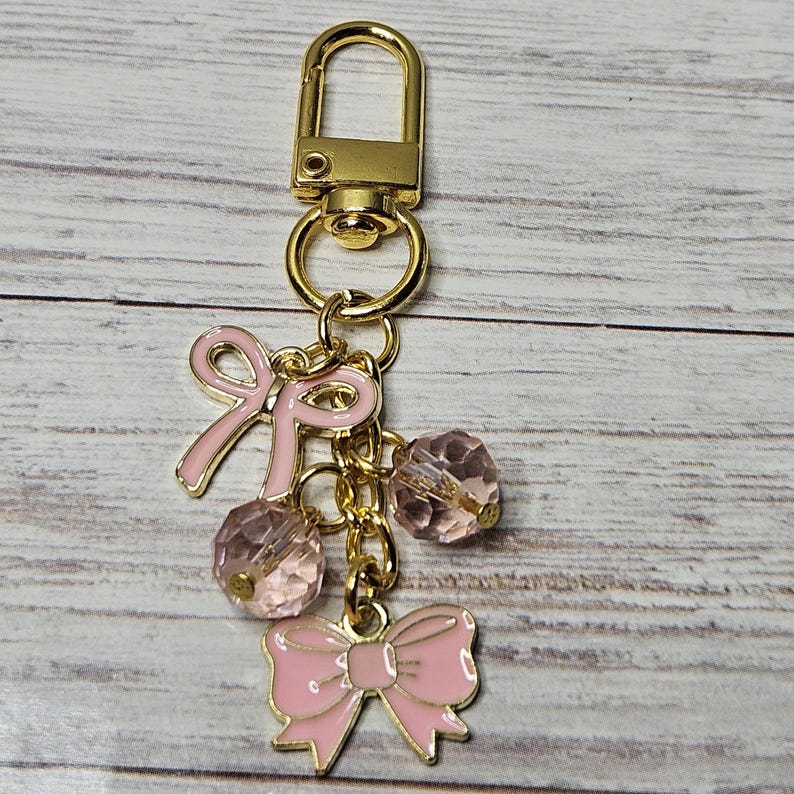 May include: A gold-colored keychain with a lobster clasp, featuring pink enamel bows and faceted pink beads. The keychain is suspended from a gold chain, creating a charming accessory.