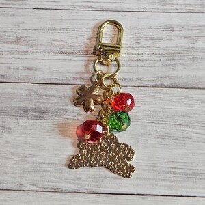 May include: Gold-toned keychain with a lobster clasp, flower charm, and a bunny-shaped charm. The keychain also features red and green faceted beads. The bunny charm has a textured pattern.