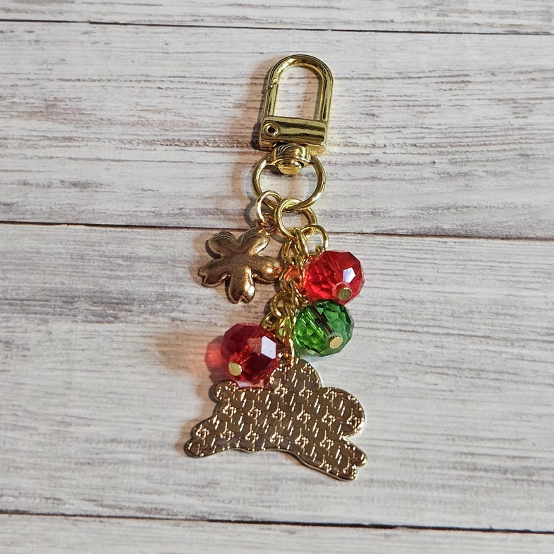 May include: Gold-toned keychain with a lobster clasp, flower charm, and a bunny-shaped charm. The keychain also features red and green faceted beads. The bunny charm has a textured pattern.