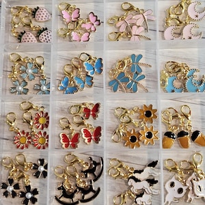 May include: A clear plastic organizer filled with various gold-toned charm pendants. The charms feature designs such as strawberries, butterflies, dragonflies, flowers, moons, and acorns. Each charm has a lobster clasp for easy attachment.
