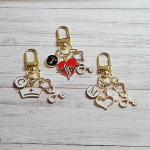 May include: Three gold-colored keychains with medical-themed charms. Each keychain has a lobster clasp and features charms like a stethoscope, a heart with a heartbeat, a nurse's cap, and letters. The charms are in red, white, and gold.