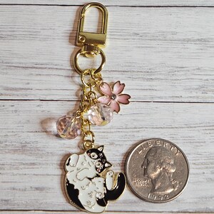 May include: A gold-colored keychain with a black and white cat charm, pink flower, and clear beads. The cat charm is lying on its back. A quarter is next to the keychain for size comparison.