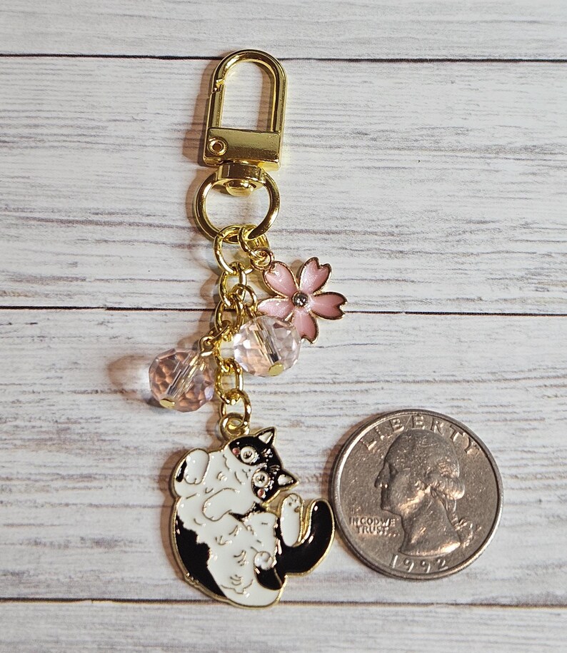 May include: A gold-colored keychain with a black and white cat charm, pink flower, and clear beads. The cat charm is lying on its back. A quarter is next to the keychain for size comparison.