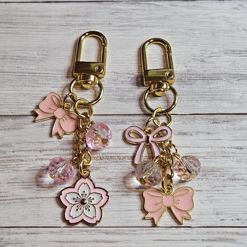 May include: Two keychains with gold-colored clasps and rings. Each keychain features pink bows, clear pink beads, and a pink and white cherry blossom charm. The keychains are hanging against a light wood grain background.