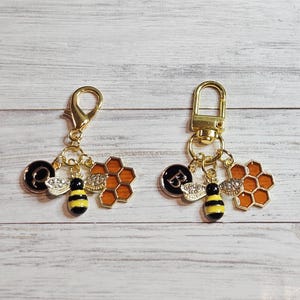 May include: Two gold-colored keychains with bee-themed charms. Each keychain features a bee, a honeycomb, and a charm with a letter. The bees are black and yellow. The honeycombs are orange.