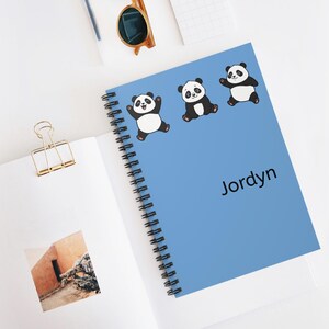 Könnte beinhalten: Ein hellblaues Spiralnotizbuch mit drei Panda-Bären-Illustrationen. Der Name "Jordyn" ist auf dem Einband aufgedruckt. Das Notizbuch ist geöffnet und zeigt weiße Seiten. Eine Sonnenbrille und eine Büroklammer sind ebenfalls zu sehen.