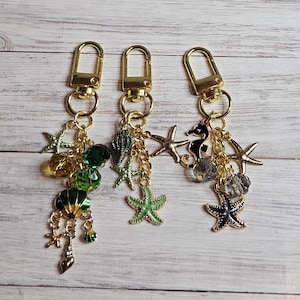 May include: Three gold keychains with marine-themed charms. Each keychain has a lobster clasp and a collection of charms, including starfish, seashells, and beads in green, gold, and black. The charms are connected by delicate gold chains.
