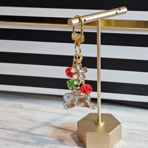 May include: A gold-colored keychain with a bunny charm, red and green beads, and a white flower charm. The bunny charm has orange floral accents. The keychain is displayed on a gold stand against a black and white striped background.