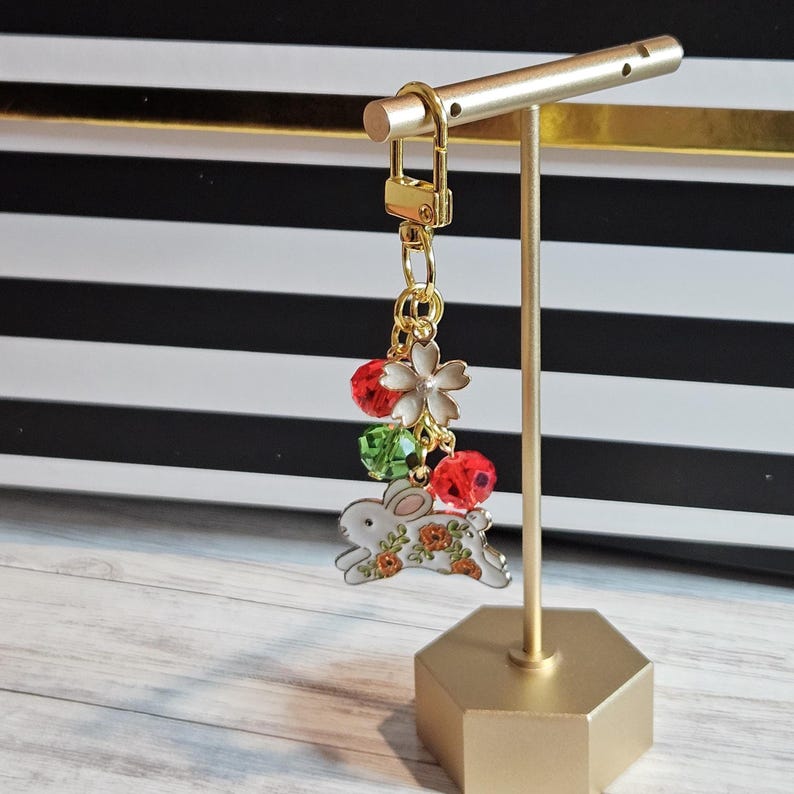 May include: A gold-colored keychain with a bunny charm, red and green beads, and a white flower charm. The bunny charm has orange floral accents. The keychain is displayed on a gold stand against a black and white striped background.