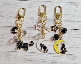 May include: Three keychains with gold-colored hardware. Each keychain features a charm with a black cat design. One has a black crescent moon, another a white crescent moon, and the last a yellow crescent moon. Each has star and bead accents.