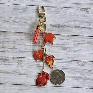 Op de afbeelding: Een goudkleurige sleutelhanger met een rode kwast en Thanksgiving-thema bedels. Bedels zijn onder meer esdoornbladeren, een kalkoen en een bedel met de tekst "GIVE THANKS". Een munt is inbegrepen ter referentie van de grootte.