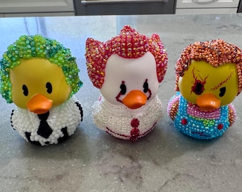 May include: Three rubber ducks decorated with colorful rhinestones. The left duck has green hair and a black tie. The center duck is white with red hair and a red tie. The right duck has orange hair, a scarred face, and blue overalls.
