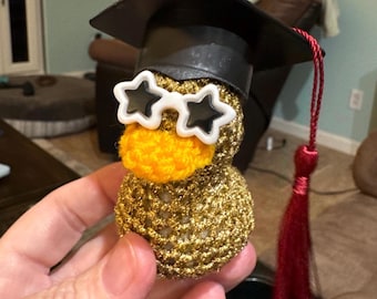 May include: A gold-colored, crocheted duck figurine wearing a black graduation cap with a red tassel and star-shaped sunglasses. The duck has an orange beak and is held in a person's hand. The figurine is a graduation-themed item.