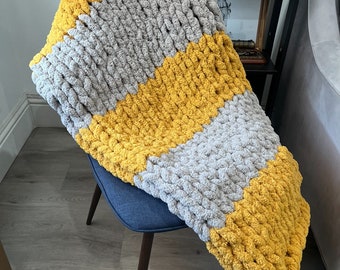 May include: A chunky knit throw blanket with stripes of mustard yellow and light gray. The blanket is draped over a chair.