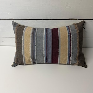 Puede incluir: Cojín decorativo rectangular con rayas verticales en tonos marrón, beige, gris, azul y burdeos. El cojín tiene un borde texturizado y deshilachado y una parte trasera marrón lisa. Para la decoración del hogar.