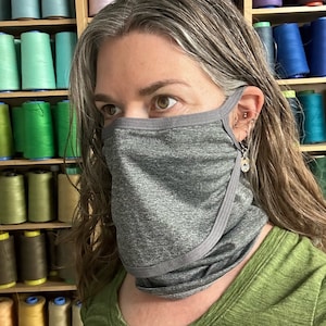 Op de afbeelding: Een persoon draagt ​​een grijs gezichtsmasker met een grijs-witte rand. Het masker is gemaakt van een zachte, ademende stof en heeft een comfortabele pasvorm. De persoon staat voor een muur met kleurrijke spoelen garen.