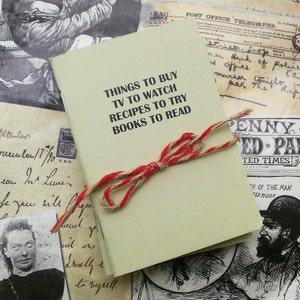 May include: A small, cream-colored notebook with a red and brown twine bow. The notebook has black text that reads "Things to Buy, TV to Watch, Recipes to Try, Books to Read".
