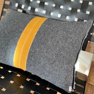 Op de afbeelding: Een grijs kussen met een gele streep in het midden. Het kussen heeft een rafelige rand en ligt op een zwart-witte gevlochten stoel.