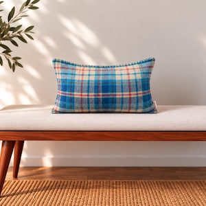 May include: A rectangular throw pillow featuring a blue, cream, and red plaid pattern. The pillow has a textured fabric surface and is finished with a decorative braided trim along the edges, providing a structured look for home decor arrangements.