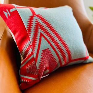 Pode incluir: Uma almofada decorativa quadrada com um padrão geométrico vermelho e azul claro. A almofada está sobre uma poltrona de couro castanho. A almofada tem uma aparência macia e texturizada.