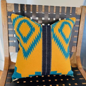 Puede incluir: Una almohada amarilla con un patrón geométrico de color verde azulado y negro. La almohada está sentada en una silla de madera con un asiento tejido negro.