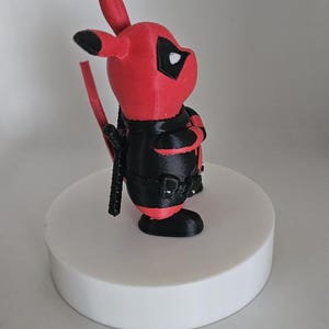 Puede incluir: Una figura impresa en 3D de color rojo y negro de un personaje Pok&eacute;mon, Pikachu, vestido como Deadpool. La figura est&aacute; de pie sobre una base circular blanca.