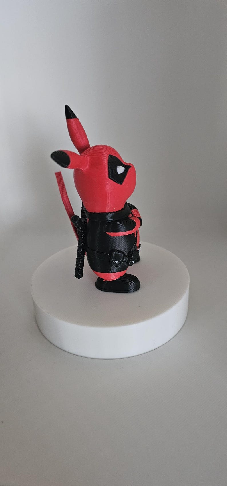 Puede incluir: Una figura impresa en 3D de color rojo y negro de un personaje Pok&eacute;mon, Pikachu, vestido como Deadpool. La figura est&aacute; de pie sobre una base circular blanca.