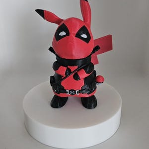 Könnte beinhalten: Eine rot-schwarze 3D-gedruckte Figur eines Pikachu, das ein Deadpool-Kostüm trägt. Die Figur steht auf einem weißen runden Sockel.