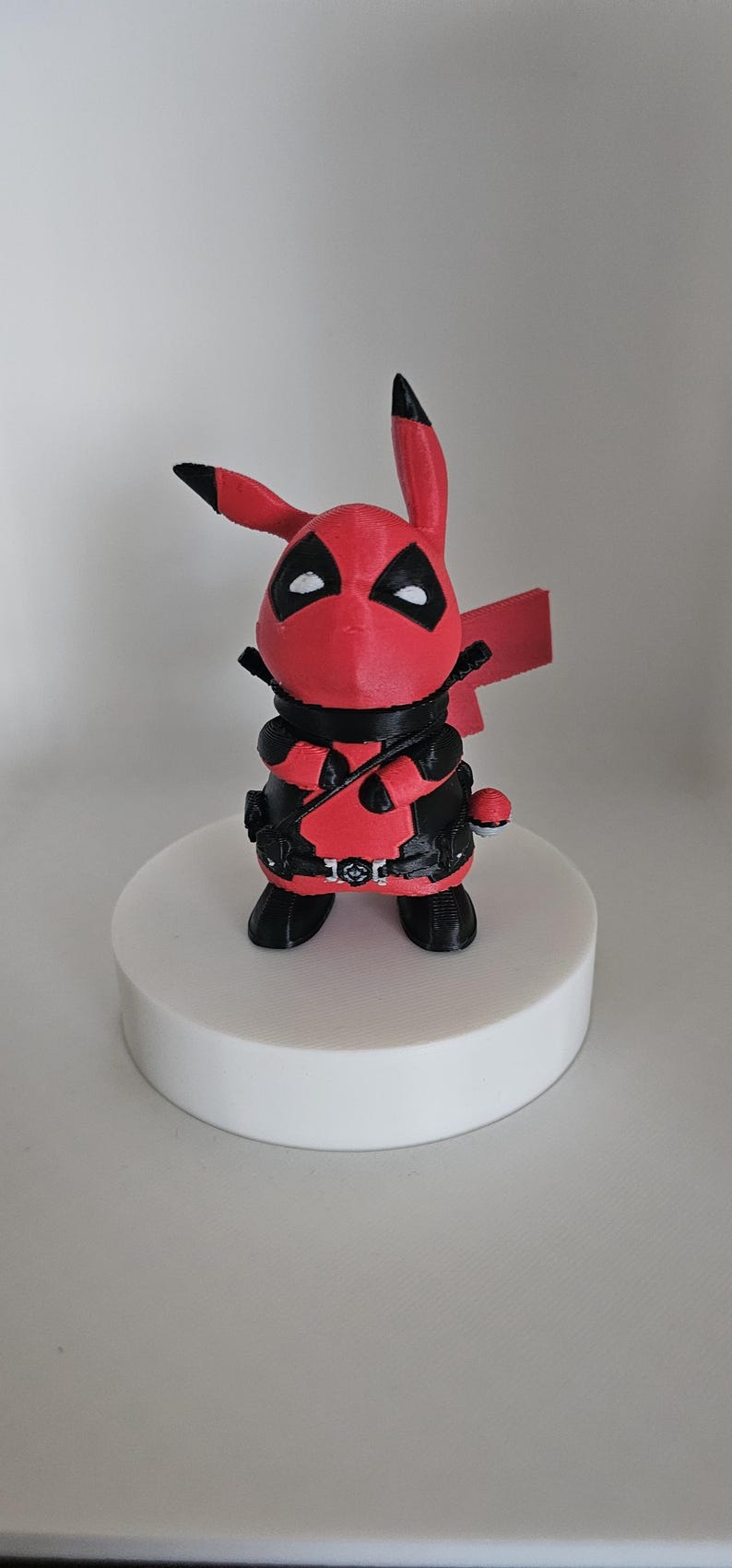 Puede incluir: Una figura impresa en 3D de color rojo y negro de un Pikachu con un disfraz de Deadpool. La figura est&aacute; de pie sobre una base circular blanca.
