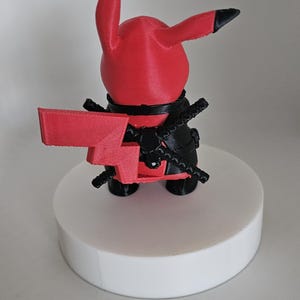 Puede incluir: Una figura de Pokemon impresa en 3D de color rojo y negro. El personaje sostiene una espada roja en su mano derecha y tiene un cintur&oacute;n negro alrededor de su cintura.