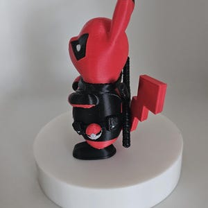 Puede incluir: Una figura impresa en 3D de color rojo y negro de un personaje Pok&eacute;mon, Pikachu, vestido como Deadpool. La figura est&aacute; de pie sobre una base circular blanca.