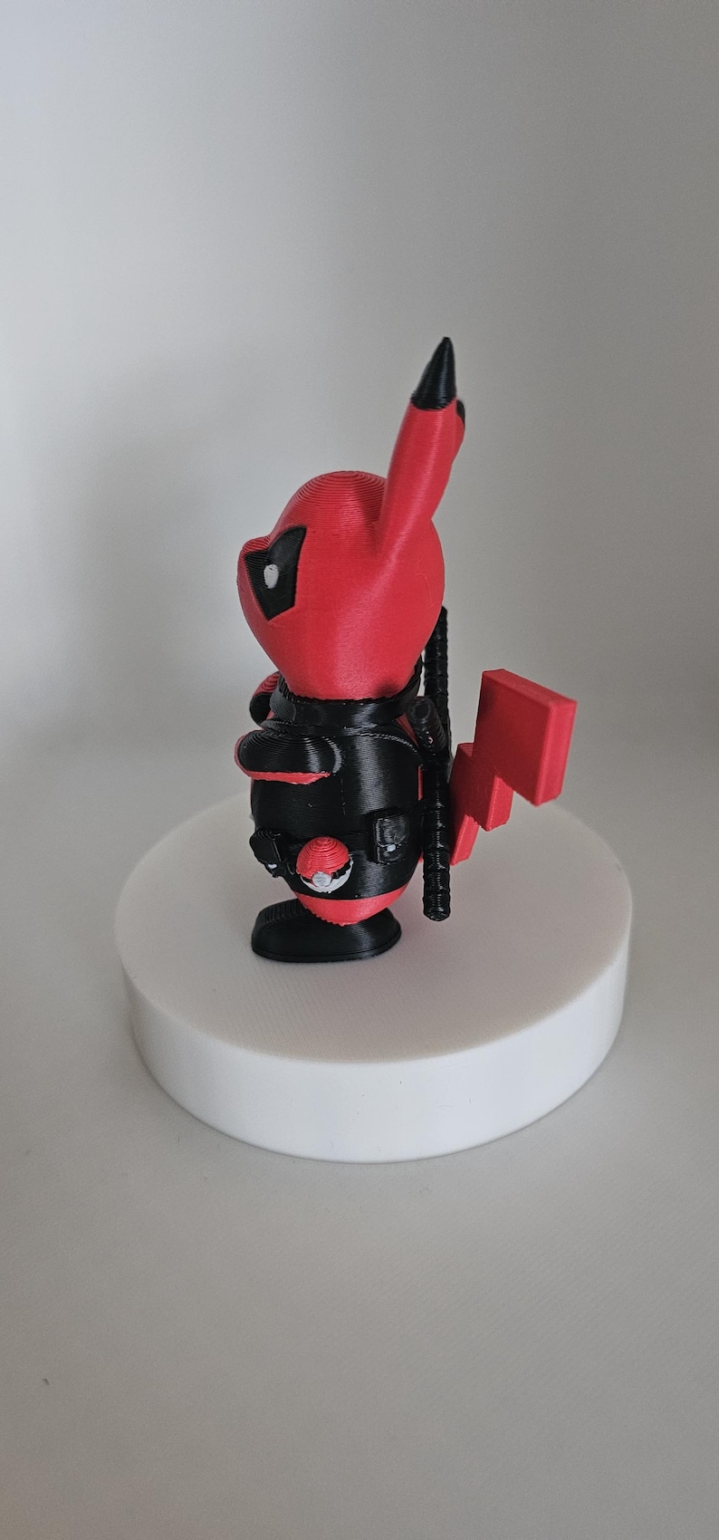 Puede incluir: Una figura impresa en 3D de color rojo y negro de un personaje Pok&eacute;mon, Pikachu, vestido como Deadpool. La figura est&aacute; de pie sobre una base circular blanca.