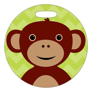 May include: A round, light green object with a handle cutout at the top, featuring a cartoon monkey face. The monkey has brown fur, tan ears and muzzle, and black eyes. The background has a green chevron pattern.